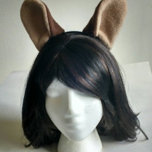 May include: A brown and tan headband with fuzzy ears. The headband is worn on a mannequin with black hair.