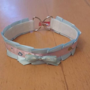 Puede incluir: Un collar de cinta azul claro y rosa con un lazo en el centro. El collar tiene una banda elástica blanca y un cierre plateado.