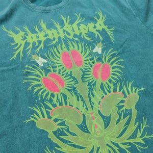 Puede incluir: Camiseta de color turquesa con un diseño gráfico de una Venus atrapamoscas con detalles rosas y verdes, y la palabra "Rooted" en una fuente estilizada. También se incluyen dos pequeñas moscas blancas en el diseño.