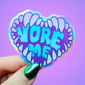 May include: A heart-shaped sticker with the words "VORE ME" in teal letters, surrounded by white teeth on a purple background. The sticker has a holographic, iridescent finish. The background is a solid purple color.