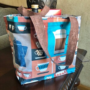 Puede incluir: Una bolsa de lona marrón con temática de café. La bolsa presenta un diseño azul y blanco con tazas de café, una cafetera y una prensa francesa. La bolsa tiene correas marrones.