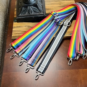 May include: A collection of colorful bag straps with silver-tone metal clasps. The straps feature rainbow, striped, and solid color designs. The straps are made of a durable, woven material.