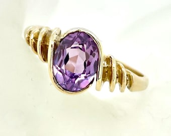 May include: A gold ring featuring an oval, light purple gemstone. The ring has a ribbed design on the sides of the band. The gemstone is set in a gold bezel, and the ring is displayed against a soft, white background.