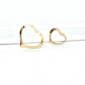 May include: Two open-heart shaped pendants in a gold tone, one larger than the other, are displayed against a white background. The pendants are simple in design, with a smooth, slightly textured surface. The heart shapes are clearly defined, and the overall aesthetic is minimalist.
