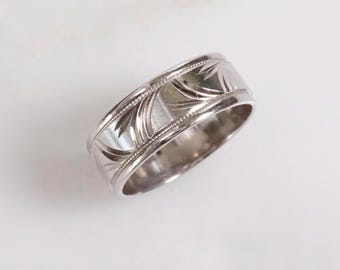 May include: A silver ring with an engraved floral pattern. The ring has a wide band with a detailed design of leaves and lines. The edges of the ring are lined with small, evenly spaced dots. The ring is shown on a white surface.