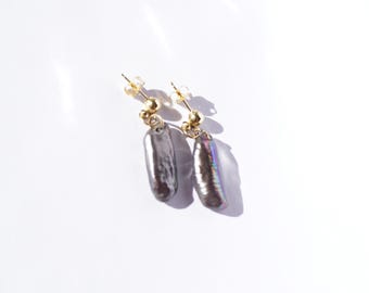 May include: A pair of drop earrings featuring elongated, irregular dark gray pearls with an iridescent sheen. Each pearl is suspended from a small, polished gold-tone metal ball stud post, creating a simple and elegant design with a metallic luster.