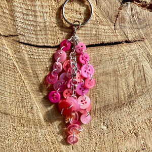 May include: A pink and white button keychain with a silver ring. The buttons are arranged in a cascading pattern, with the largest buttons at the top and the smallest buttons at the bottom.