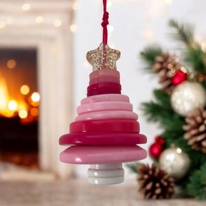May include: A pink and red Christmas tree ornament. The ornament is made of graduated circles, topped with a star. A red string is attached for hanging. The background includes a Christmas tree and a fireplace. The ornament is designed for festive decoration.