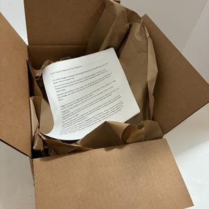 May include: An open cardboard box with brown packing paper and a printed document inside. The document has black text on a white background. The box is on a white surface.