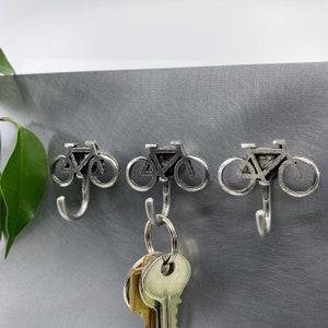 May include: Three silver bicycle-shaped hooks mounted on a gray wall. A set of keys is hanging on one of the hooks.