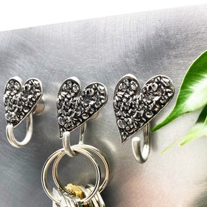 May include: Three silver heart-shaped hooks mounted on a silver metal surface. The hooks have a textured design and are ideal for hanging keys or other small items.