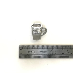 May include: A silver-coloured metal mug with a leaf design. The mug is approximately 2.5 cm tall and is sitting on a ruler.