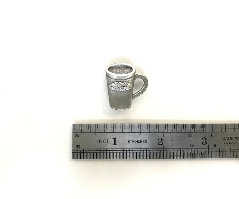 May include: A silver-coloured metal mug with a leaf design. The mug is approximately 2.5 cm tall and is sitting on a ruler.