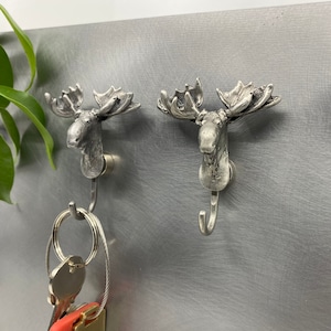 May include: Two silver-colored moose head hooks with detailed antlers are mounted on a metallic surface. One hook holds a set of keys. The hooks are designed for hanging items and add a rustic touch to any space.