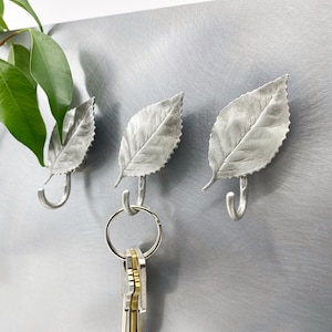 Puede incluir: Tres ganchos de plata en forma de hoja montados en una pared de plata. Un juego de llaves cuelga de uno de los ganchos.