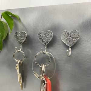 May include: Three silver heart-shaped hooks with intricate mandala designs. Each hook has a key ring attached and is holding keys.