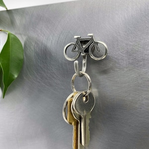 May include: A silver bicycle-shaped hook with a silver key ring and keys hanging from it. The hook is mounted on a silver wall.