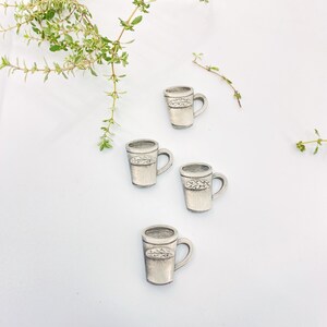 May include: Four silver-coloured ceramic mugs with a leaf design on the front. The mugs are arranged in a row on a white surface.