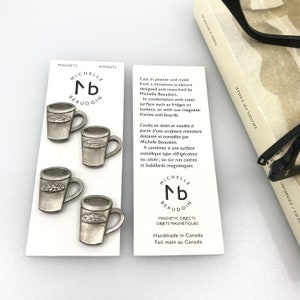 May include: Four silver-toned metal mugs with a leaf design. The mugs are magnetic and are packaged with a white card that reads "Michelle nb Beaudoin" and "Magnetic Objects, Handmade in Canada".