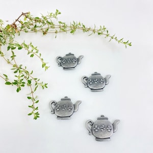 May include: Four silver teapot-shaped charms with a floral design. The charms are on a white background.