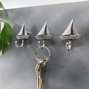 Puede incluir: Tres ganchos para llaves de plata con forma de velero montados en una superficie de metal plateado. Un juego de llaves cuelga de uno de los ganchos.