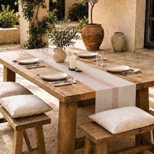 Può includere: Un lungo tavolo da pranzo in legno con due panche abbinate, ciascuna dotata di un cuscino di colore chiaro. Il tavolo è apparecchiato con un runner beige con una striscia rossa centrale, piatti bianchi, bicchieri trasparenti e un vaso.