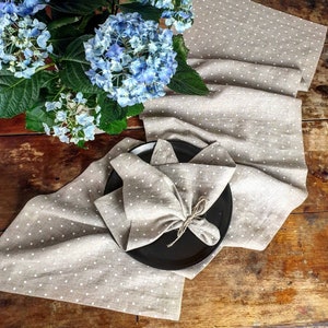 Puede incluir: Un camino de mesa de lino beige con lunares blancos, doblado en una servilleta decorativa sobre un plato negro. Un ramo de hortensias azules está en el fondo.