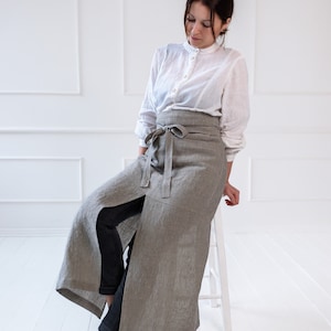 May include: A woman wearing a white button-down shirt and a long, striped linen apron with a tie waist. She is sitting on a white stool with her legs crossed.