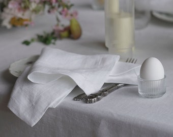 May include: A white egg in a clear glass egg cup on a white tablecloth with a white linen napkin and a silver fork. There are flowers in the background.