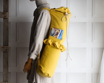Puede incluir: Una bolsa de yoga de lino amarilla con un borde con volantes y un pequeño bolsillo. La bolsa tiene un cierre de cordón y correas ajustables.