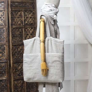 May include: A large, light gray linen tote bag with white crocheted trim. The bag is being held by a wooden hand on a mannequin.