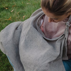 Pu&ograve; includere: Una donna che indossa un maglione rosa e una copertina per l'allattamento a righe sta allattando al seno un bambino. La copertina &egrave; realizzata in un tessuto morbido e traspirante ed &egrave; progettata per fornire privacy e comfort sia alla madre che al bambino.