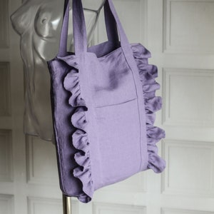 Pode incluir: Uma bolsa tote de linho lavanda com bordas de babados e um único bolso na frente.