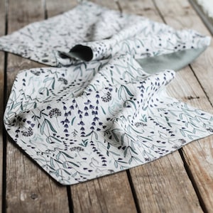 May include: A white linen table runner with a green and purple floral pattern. The runner is folded and lying on a wooden surface.