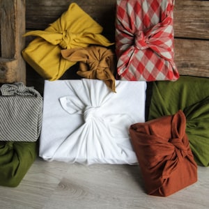 May include: A collection of six gifts wrapped in various fabrics, including a white fabric, a red and white checkered fabric, a yellow fabric, a brown fabric, a green fabric, and a striped fabric.