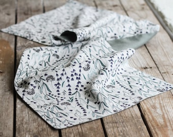 May include: A white linen table runner with a green and purple floral pattern. The runner is folded and lying on a wooden surface.