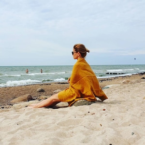 May include: A woman wearing sunglasses sits on a sandy beach, wrapped in a mustard yellow towel, looking out at the ocean.