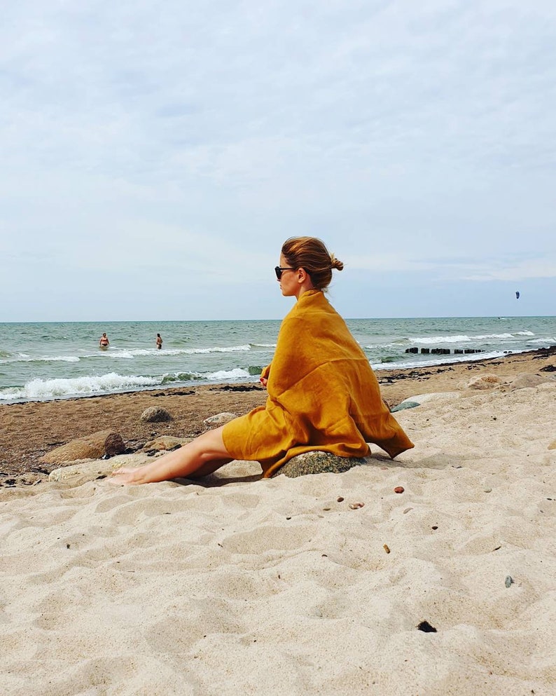 May include: A woman wearing sunglasses sits on a sandy beach, wrapped in a mustard yellow towel, looking out at the ocean.