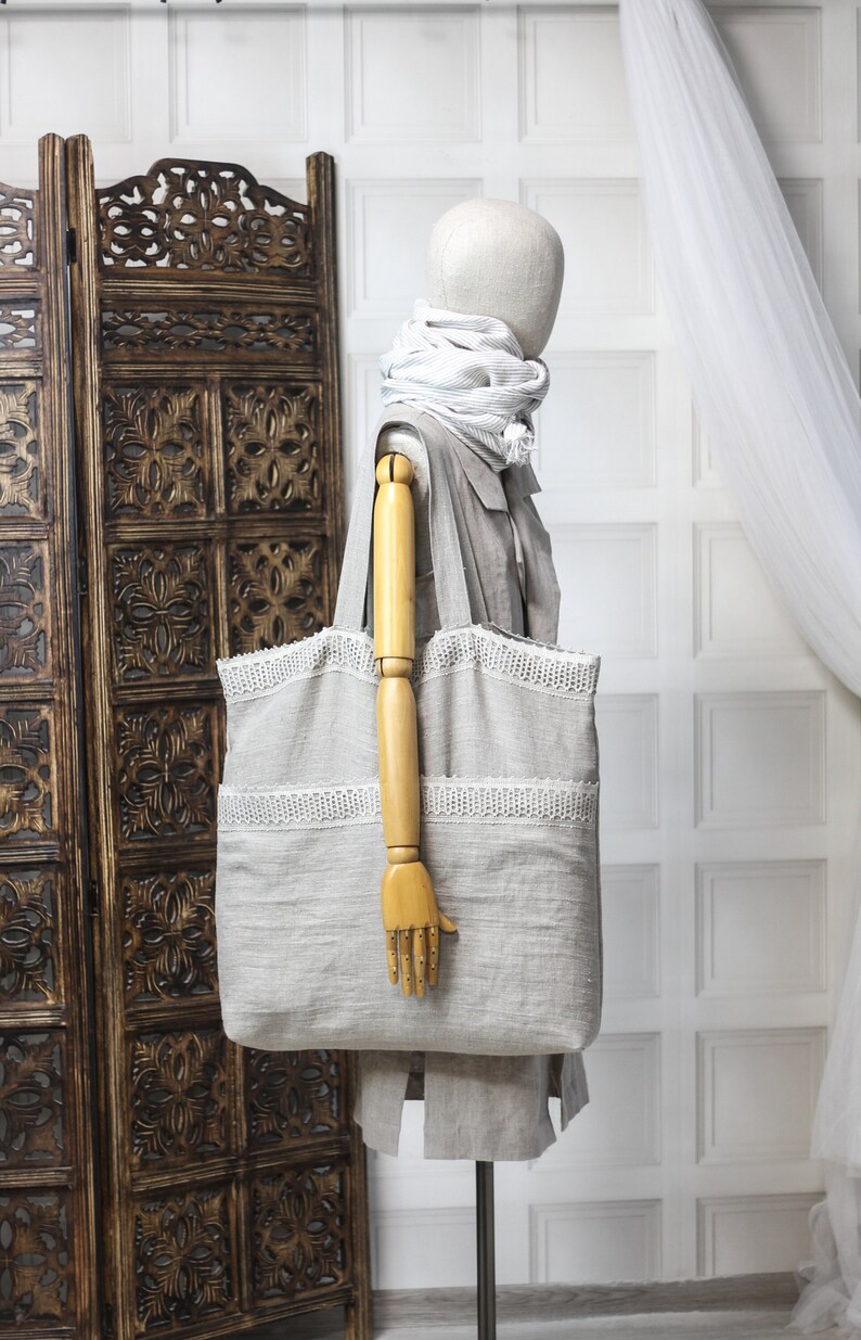 May include: A large, light gray linen tote bag with white crocheted trim. The bag is being held by a wooden hand on a mannequin.