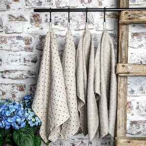 May include: Four beige linen tea towels with black polka dots hanging on a black metal rod. The towels are hanging in a kitchen or bathroom setting.