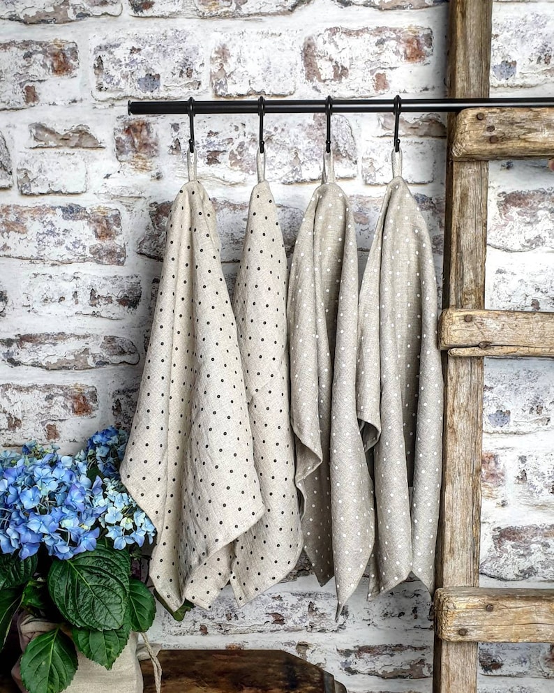 May include: Four beige linen tea towels with black polka dots hanging on a black metal rod. The towels are hanging in a kitchen or bathroom setting.