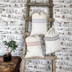 Op de afbeelding: Drie linnen trekzakken met gestreepte ontwerpen. De zakken hangen aan een houten ladder tegen een witte bakstenen muur.