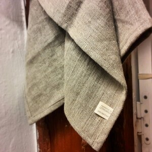 May include: A beige, textured fabric towel featuring a visible weave and a small white rectangular label sewn onto the corner. The label contains the black printed text: HANDMADE IN LITHUANIA LOVELY CRAFTS HOME. The fabric appears folded and draped.