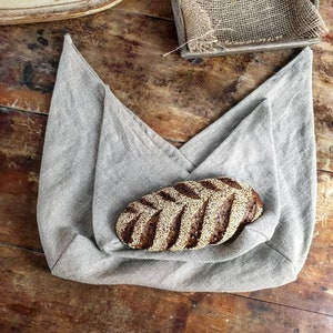 May include: A grey linen bag with a loaf of dark brown bread with sesame seeds inside.