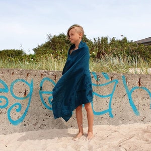 May include: A young person standing on a sandy beach wearing a teal blue towel wrapped around them. The person is standing in front of a concrete wall with blue graffiti.