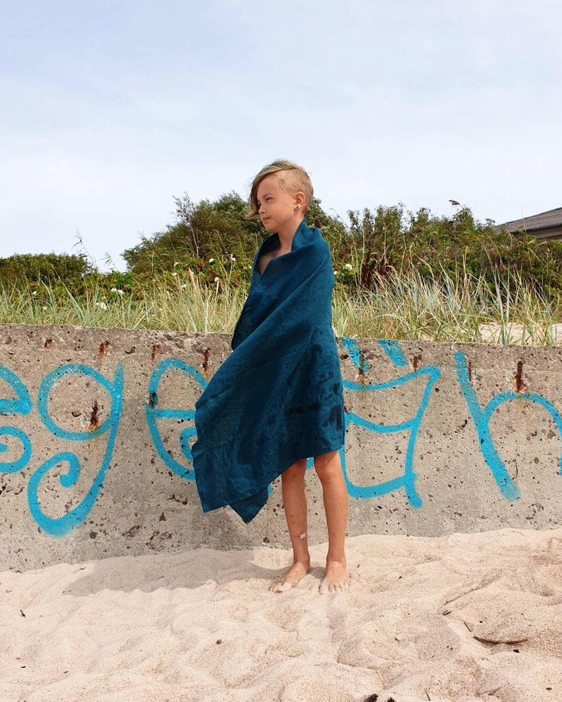 May include: A young person standing on a sandy beach wearing a teal blue towel wrapped around them. The person is standing in front of a concrete wall with blue graffiti.