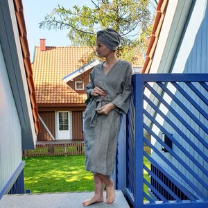 May include: A woman wearing a gray striped robe and a gray turban stands on a wooden deck with a blue railing. The background shows a red-roofed house with a green lawn.