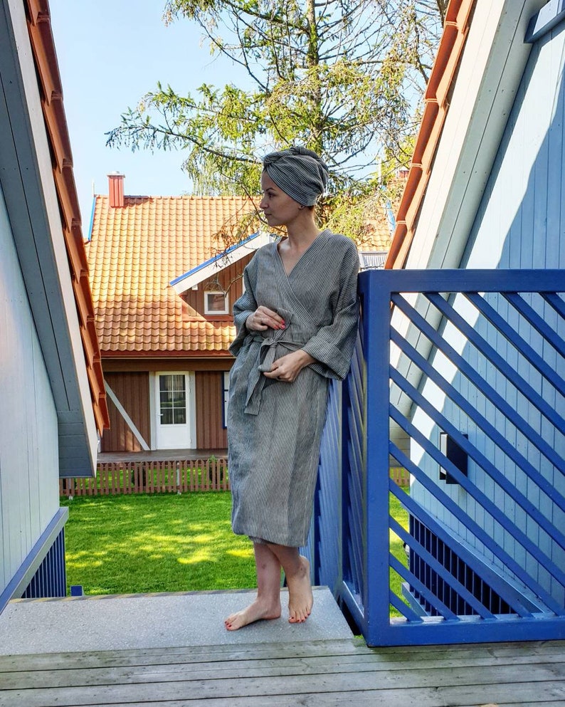 May include: A woman wearing a gray striped robe and a gray turban stands on a wooden deck with a blue railing. The background shows a red-roofed house with a green lawn.