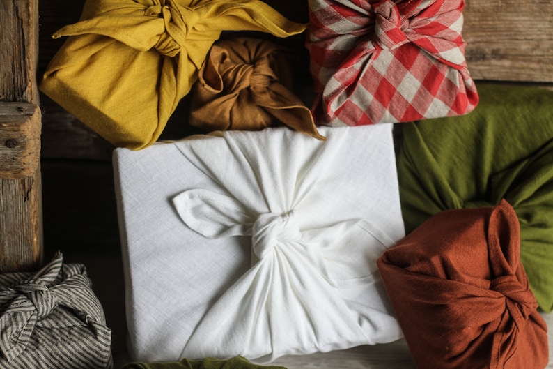 May include: A collection of gifts wrapped in various fabrics, including a white fabric, a yellow fabric, a brown fabric, a red and white checkered fabric, a green fabric, and a brown fabric. The gifts are tied with knots and bows.
