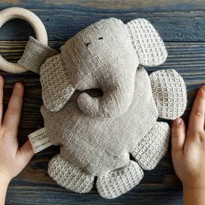 Peut inclure: Un jouet éléphant en lin fait à la main avec des oreilles et des pieds texturés en gaufre, et un anneau en bois. Le jouet est gris clair et porte une étiquette indiquant "Handmade in Lithuania".